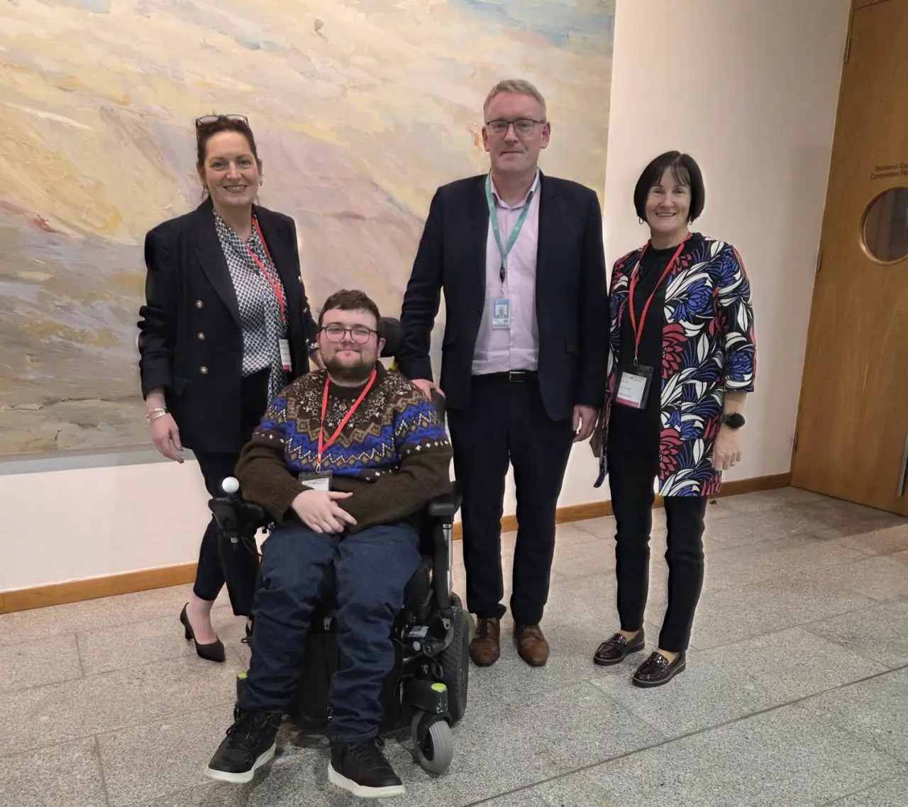 A male wheelchair user pictured with two women and one gentleman who are standing. They are in an indoor setting.
