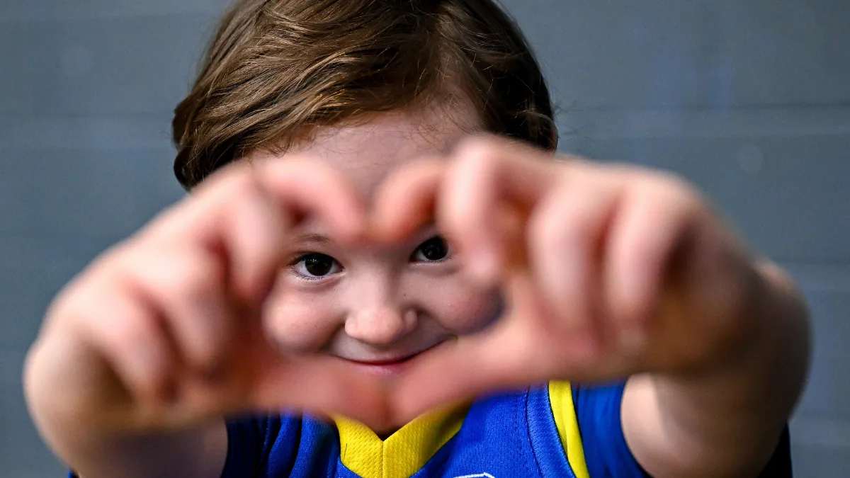A child makes a love heart with her hands while smiling. She is wearing a blue and yellow sports top