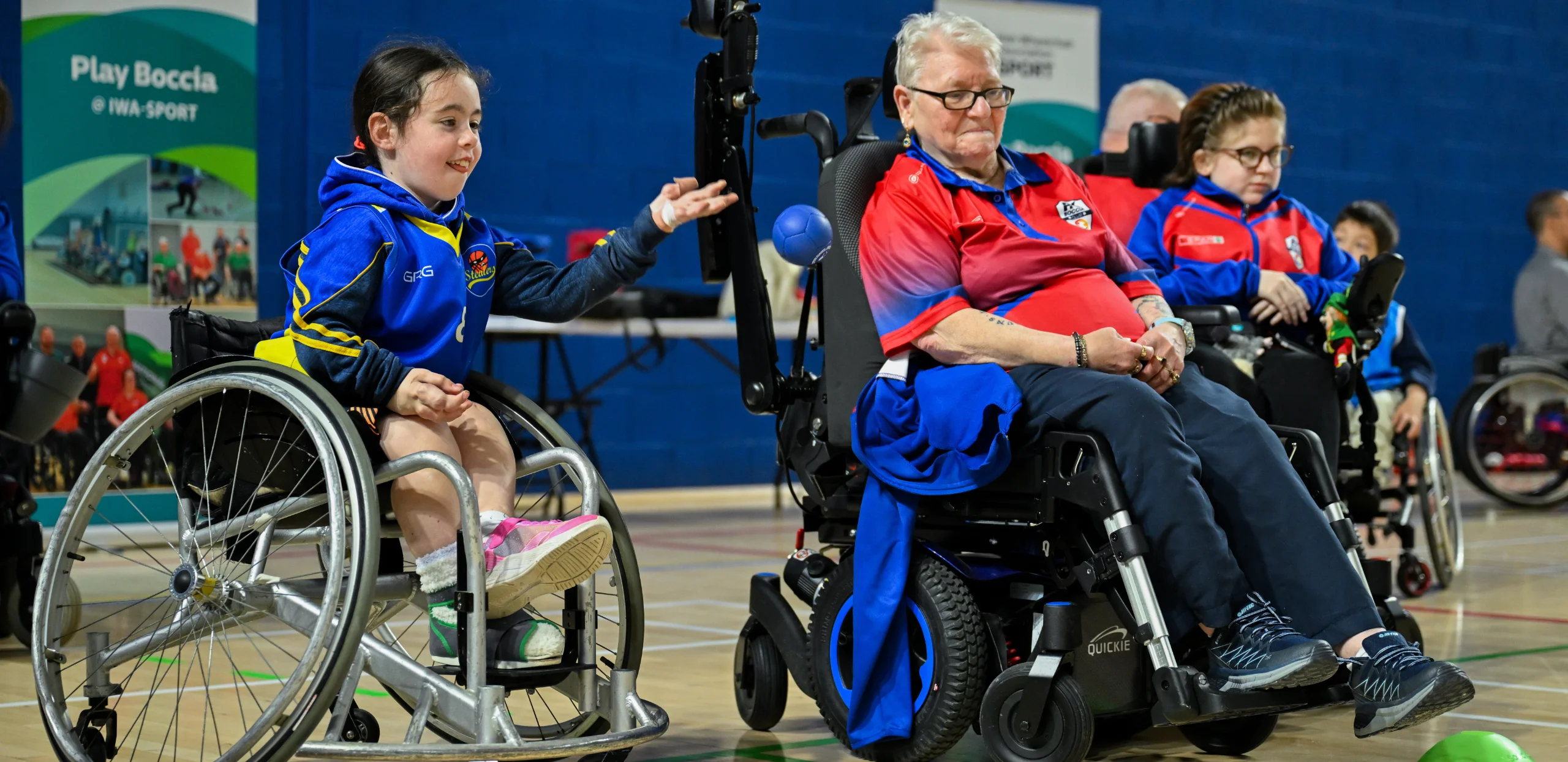A girl in a blue and yellow basketball top sits in a basketball wheelchair, left arm outstretched after throwing a blue ball, which is centre of the image. To the right is an older woman in a powered wheelchair, wearing a red and blue polo shirt and a jumper hanging from her chair, looking on. In the background to the right is a girl in a power chair playing Boccia.