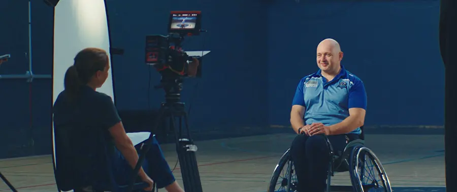 Man in a wheelchair photographed being filmed during an interview with a woman sitting off camera